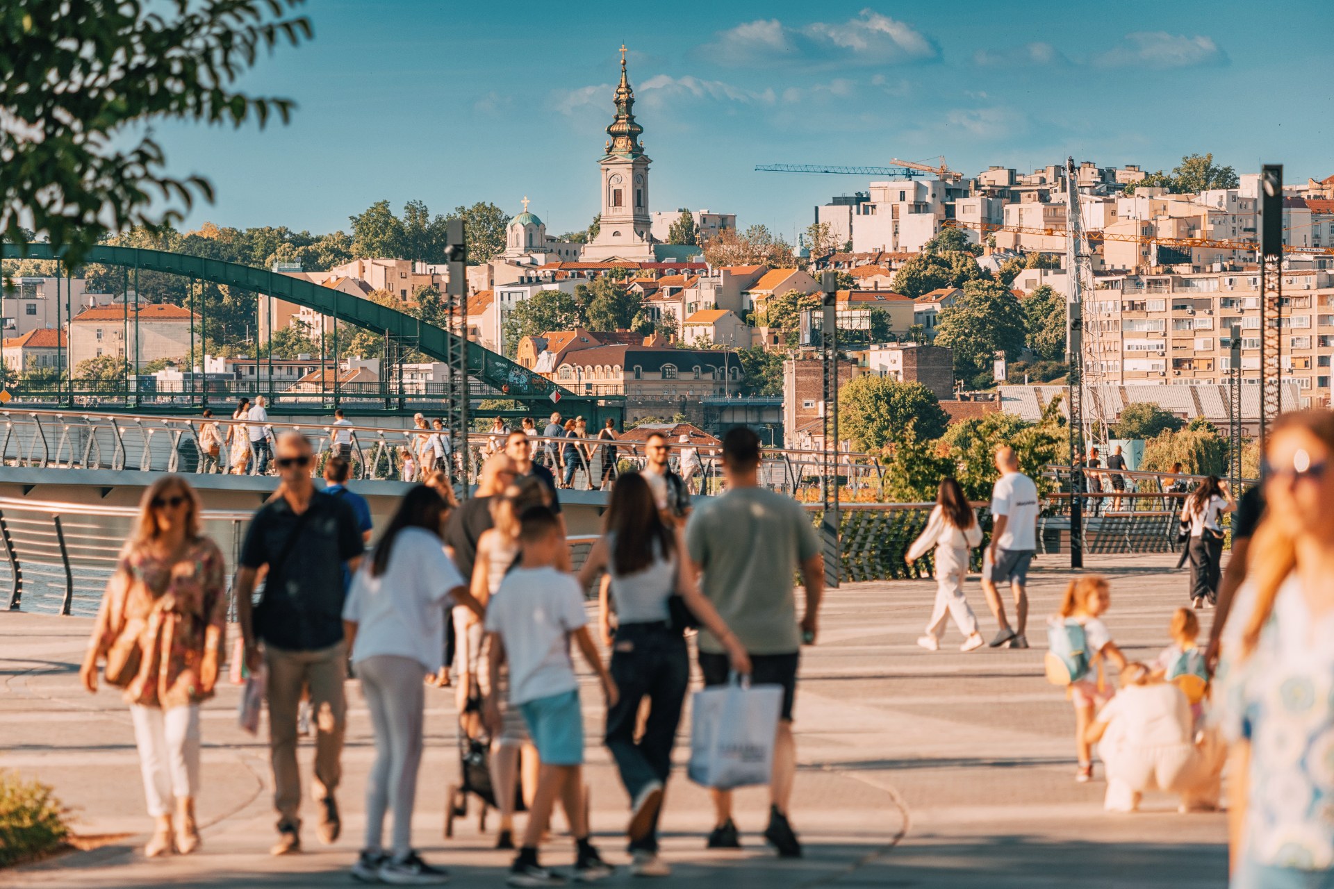 Sétálók a belgrádi Száva-parti sétányon, a háttérben az óváros és egy templomtorony
