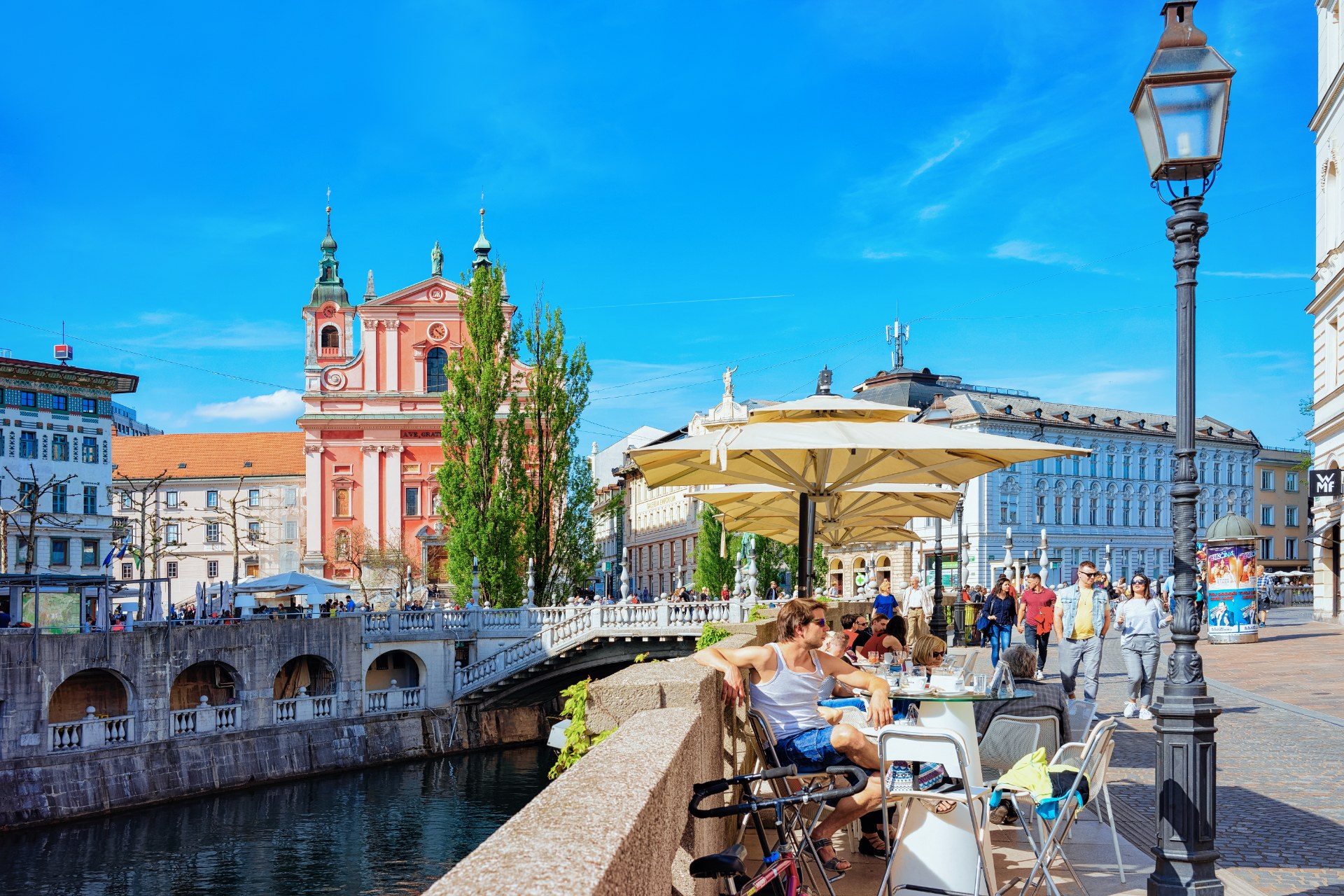 Emberek egy kávézó teraszán a ljubljanai Hármas hídnál, a Ljubljanica partján